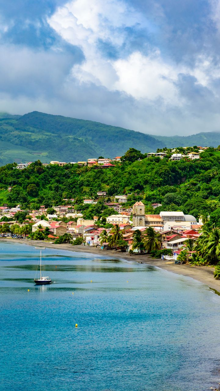Ville côtière au bord d'une mer bleue. Maisons nichées à flanc de colline verdoyante sous un ciel nuageux. Voilier sur l'eau.