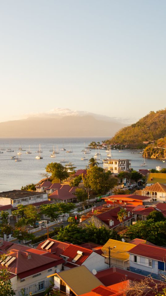 Ville côtière aux toits colorés, voiliers sur l'eau et montagnes à l'horizon.