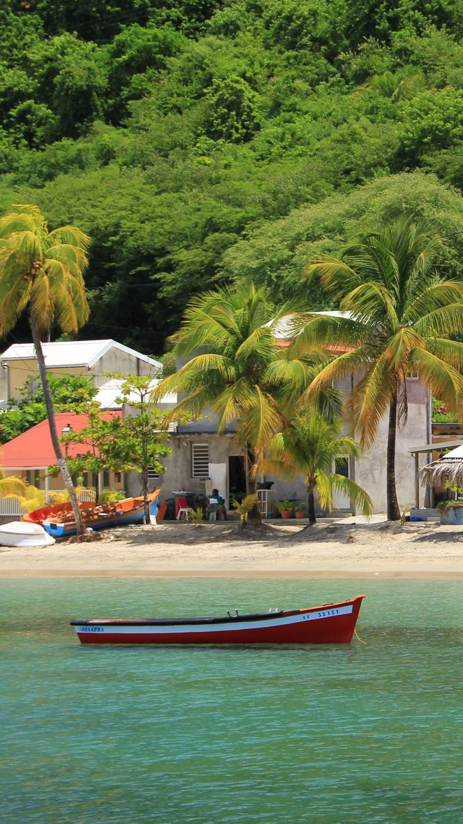 Bateau rouge dans une eau turquoise près d'une plage bordée de bâtiments et d'une végétation luxuriante.