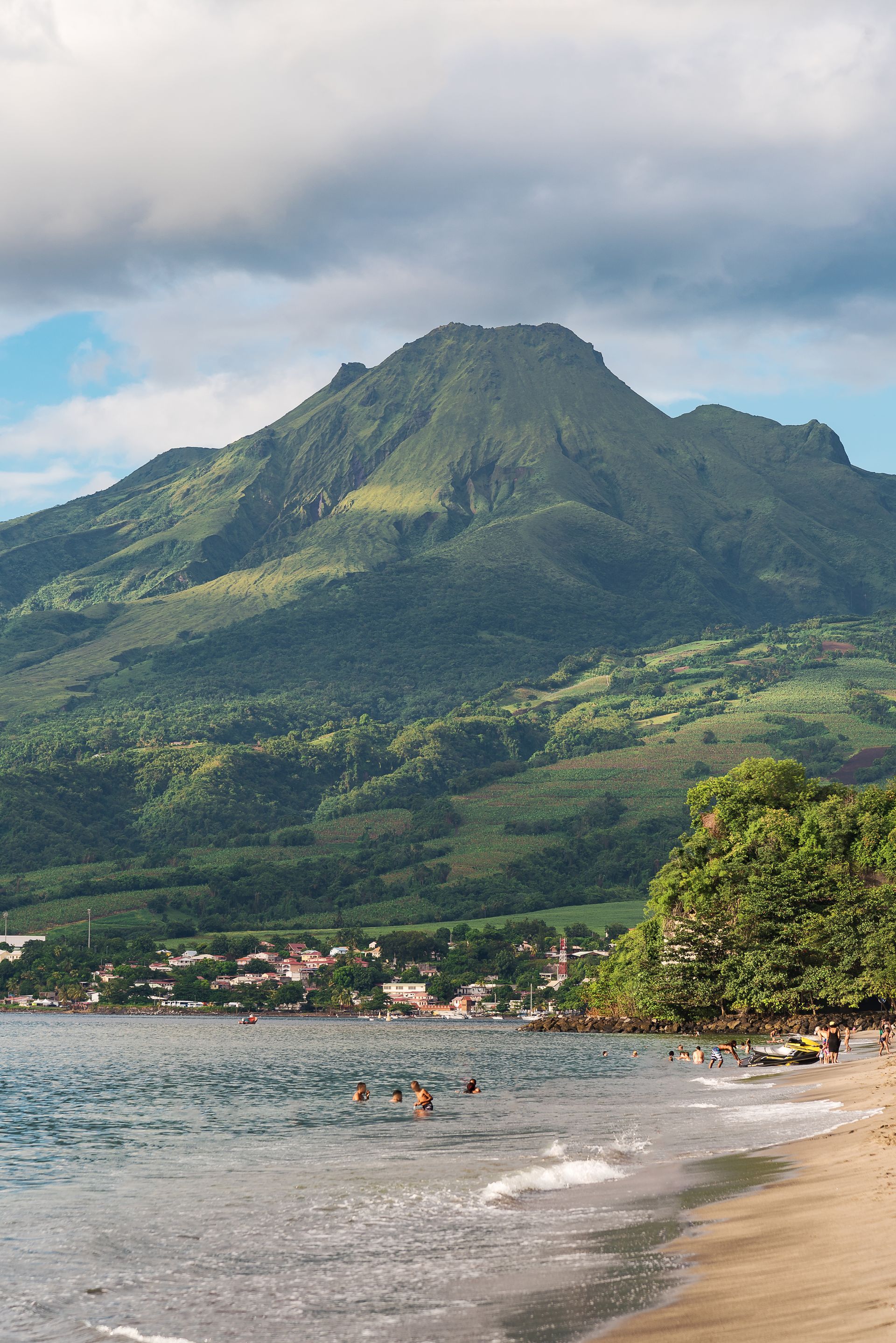 Plage avec des baigneurs, des maisons et une montagne verdoyante sous un ciel nuageux.