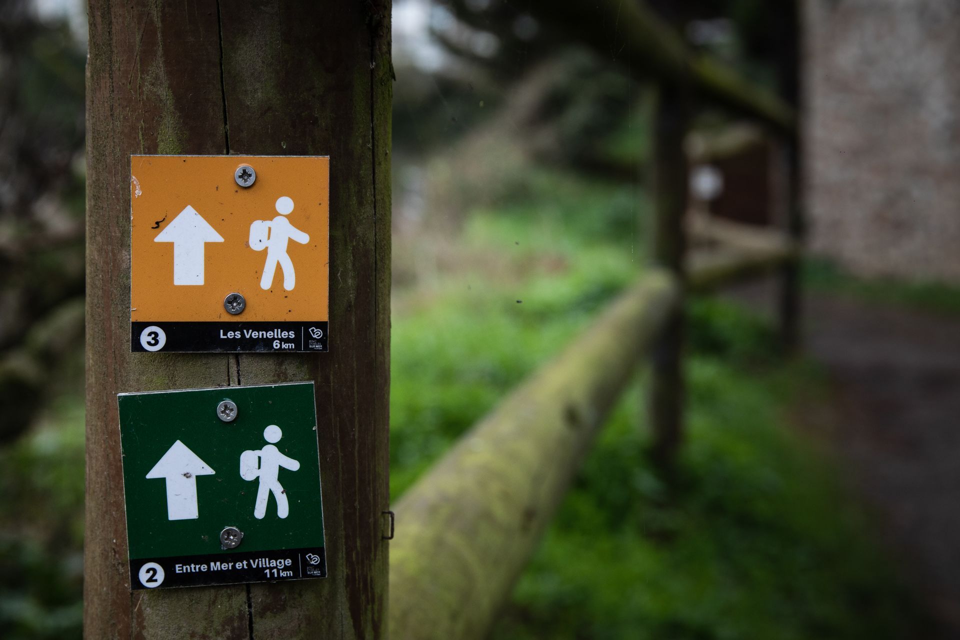 Deux panneaux de signalisation, orange et vert, avec des symboles de randonneurs et des flèches, fixés sur un poteau en bois.