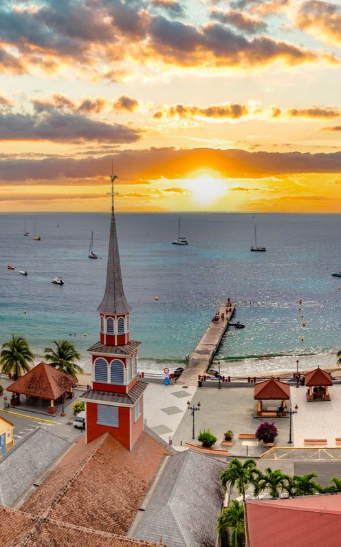 Coucher de soleil sur une ville avec des bâtiments aux toits rouges, une jetée et des voiliers sur l'eau.