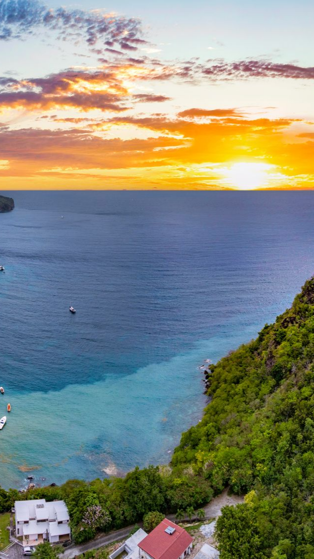 Coucher de soleil sur une baie océanique aux eaux peu profondes turquoise, aux falaises verdoyantes et aux petits bâtiments.