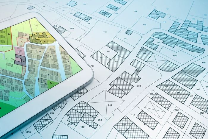 Tableta que muestra un mapa colorido sobre planos arquitectónicos en una mesa de dibujo.