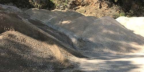 Des tas de gravier et de terre sur une surface pavée ombragée, avec une colline en arrière-plan.