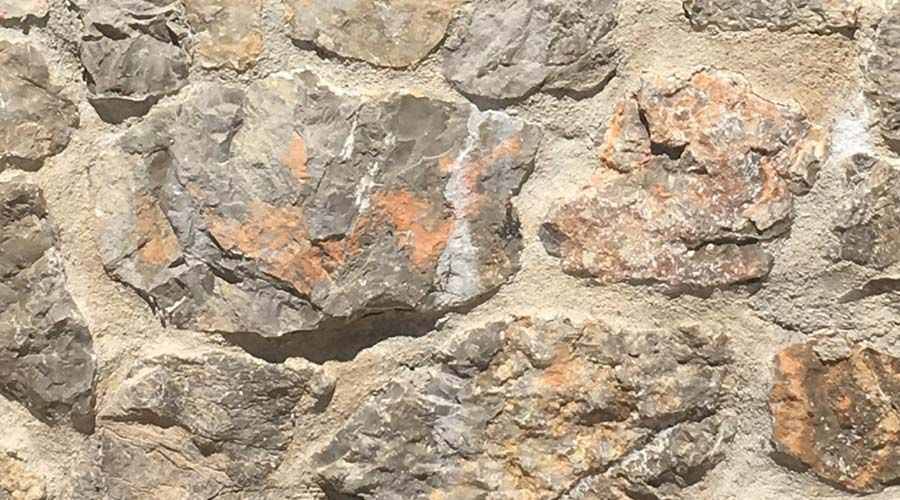 Gros plan sur un mur de pierre. Des pierres grises et beiges, taillées grossièrement, sont assemblées avec du mortier, certaines présentant des reflets orangés.