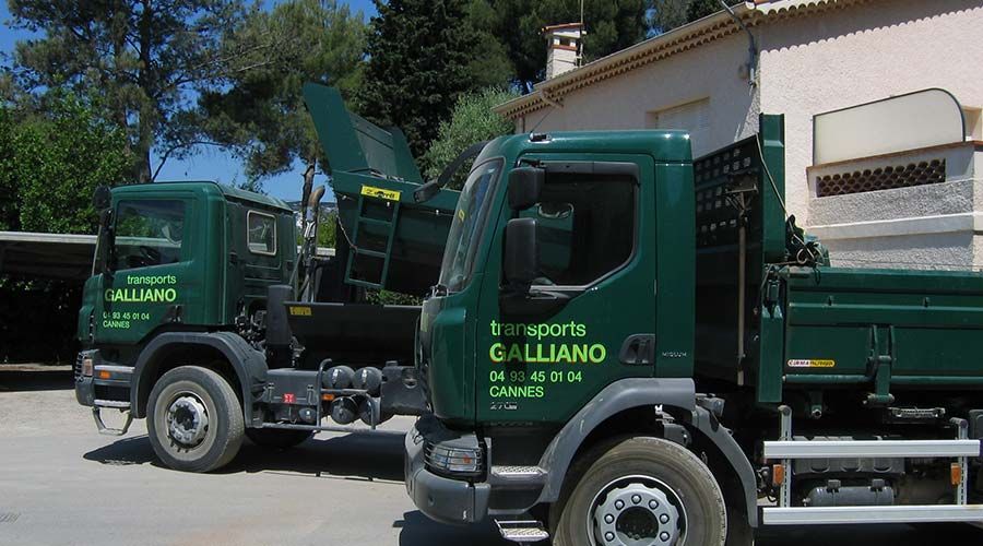 Deux camions verts « Transports Galliano » sont garés dehors. L'un d'eux est équipé d'un broyeur de branches. Journée ensoleillée.