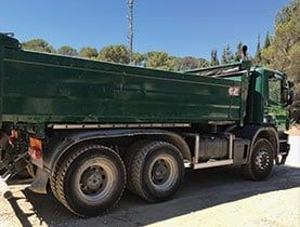 Un camion-benne vert sur un chemin de gravier, sous un ciel bleu, dans un cadre ensoleillé.