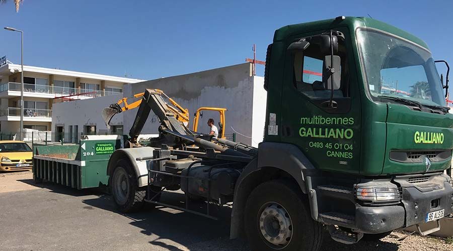Un camion Galliano vert, chargé de déchets, transporte une benne à ordures près d'un bâtiment en construction par une journée ensoleillée.
