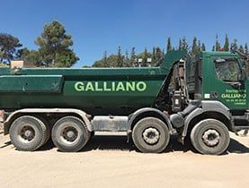 Un camion-benne Galliano vert sur une route poussiéreuse sous un ciel bleu.
