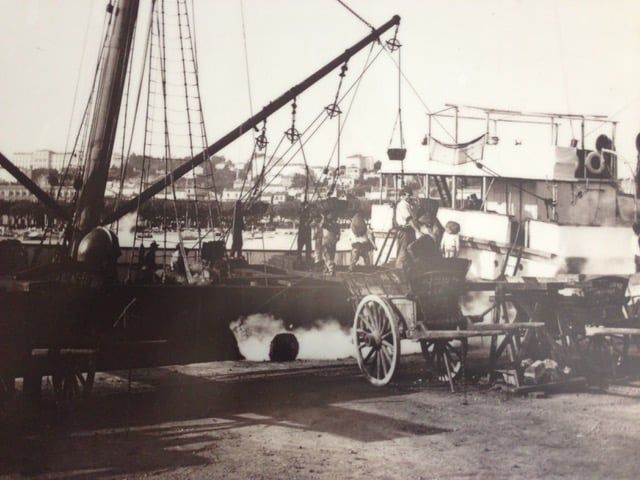 Photo en noir et blanc d'un bateau déchargeant sa cargaison dans des chariots sur un quai en présence de personnes.