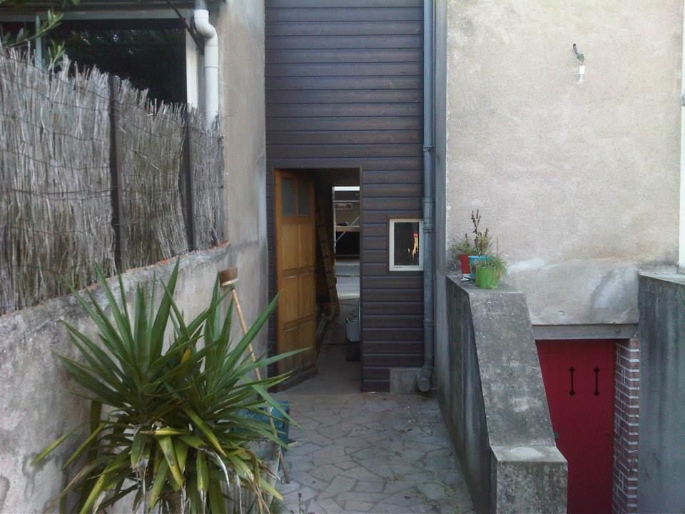 Un étroit chemin de pierre mène à une porte située à côté d'un mur beige et d'un bardage sombre, avec des plantes en pot et une rambarde.