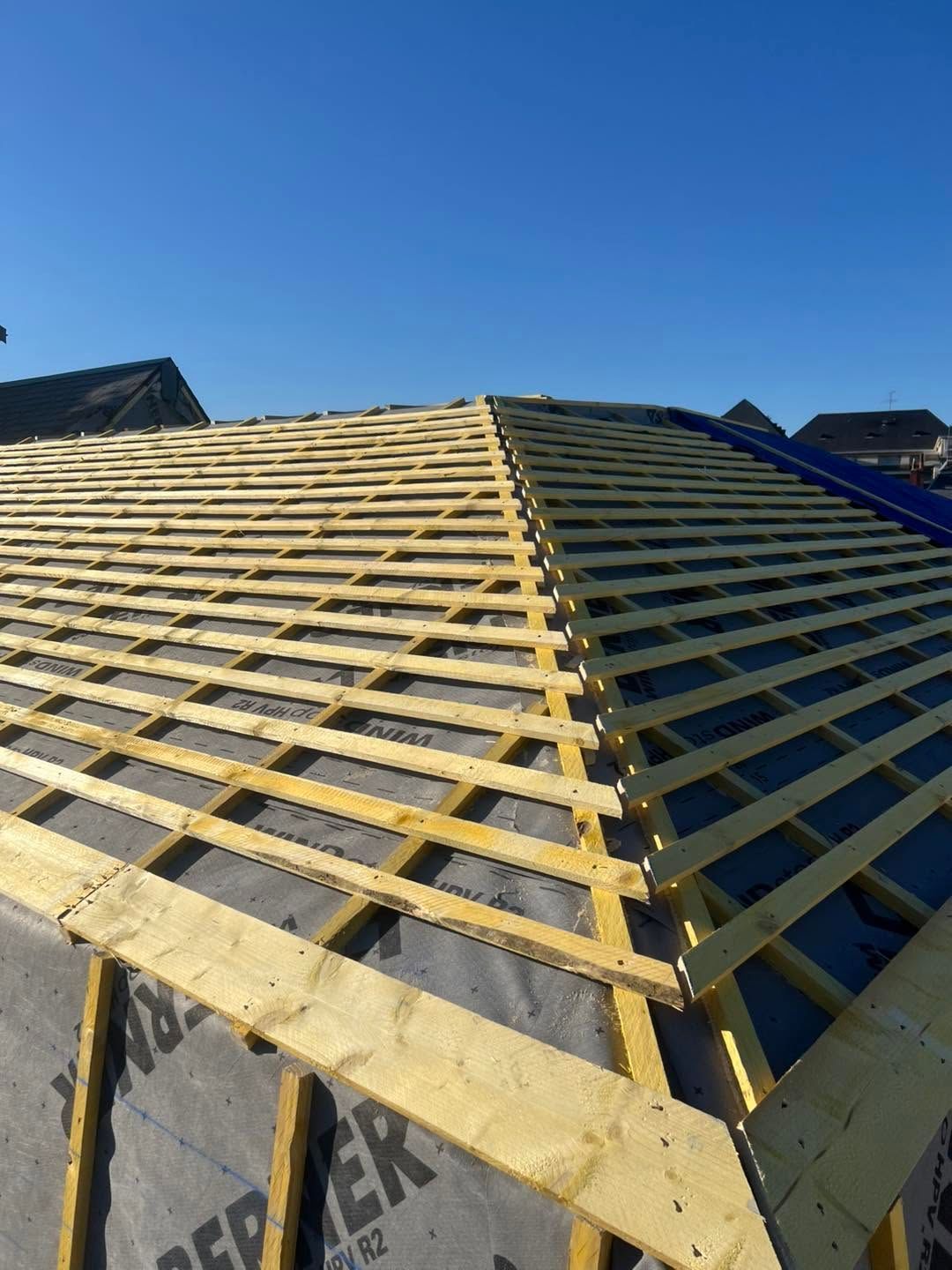 Charpente de toit en construction, avec des fermes en bois jaunes se détachant sur un ciel bleu.