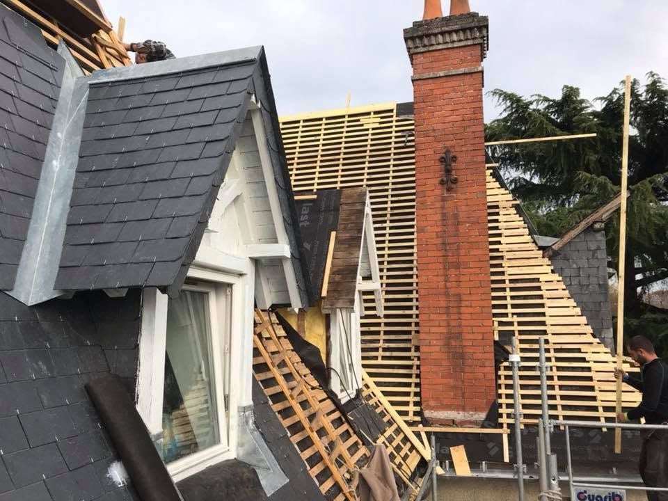 Toiture en construction avec bardeaux d'ardoise et cheminée en briques rouges, charpente apparente.