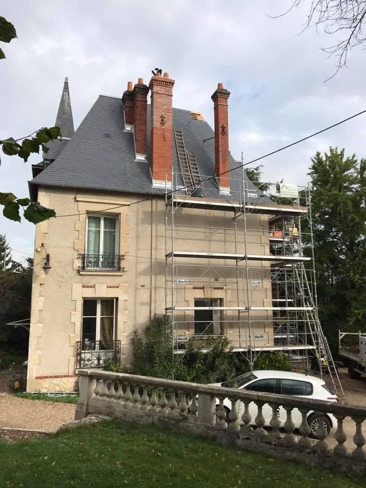 Maison en pierre avec toit d'ardoise et échafaudages, vue depuis un jardin avec une voiture garée en contrebas.