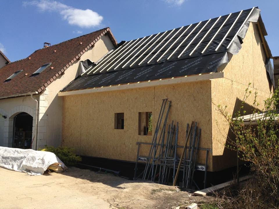 Maison en construction avec un toit noir, des échelles appuyées contre le mur et des matériaux de construction à proximité.