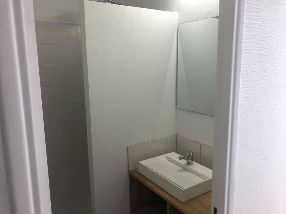 Petite salle de bains avec lavabo, miroir et porte de cabine grise, vue à travers une porte.