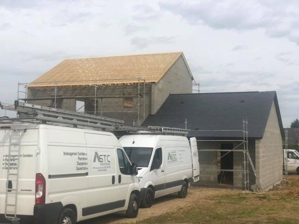 Deux fourgonnettes blanches de chantier sont garées près d'une maison en construction, avec des échafaudages et une charpente de toit apparente.