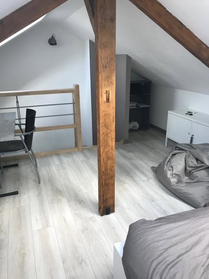 Chambre en mezzanine avec plafond blanc incliné, poutre en bois apparente, bureau, chaise, pouf et lit.