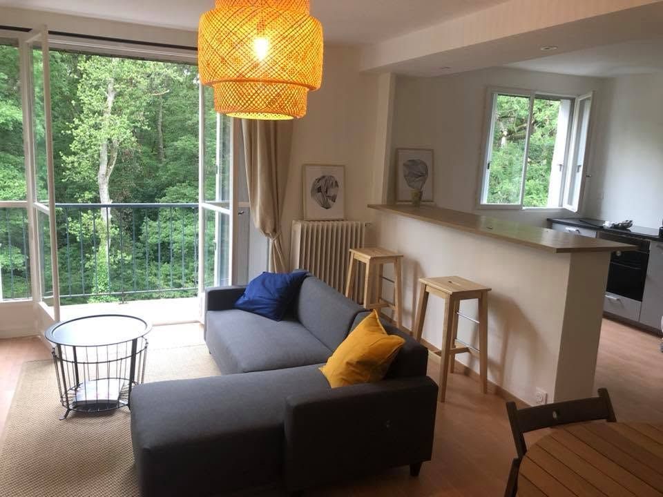 Salon avec canapé gris, lampe jaune et grandes fenêtres donnant sur la verdure.