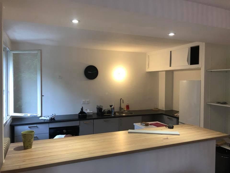 Cuisine moderne avec îlot central en bois clair, placards gris, murs blancs et petite table près d'une fenêtre.