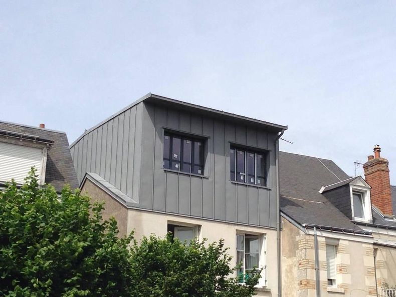 Extension moderne grise d'une maison avec des fenêtres à cadre foncé, jouxtant des maisons en pierre plus anciennes.