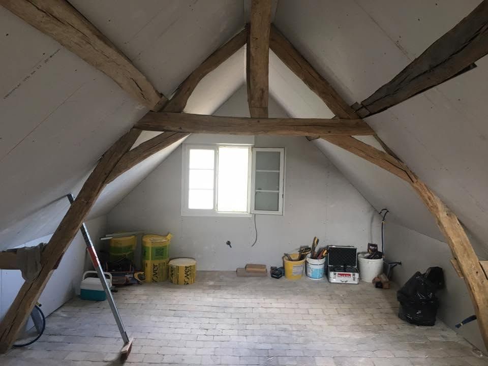 Pièce mansardée avec poutres apparentes, murs blancs, une fenêtre et outils et caisses rangés au sol.
