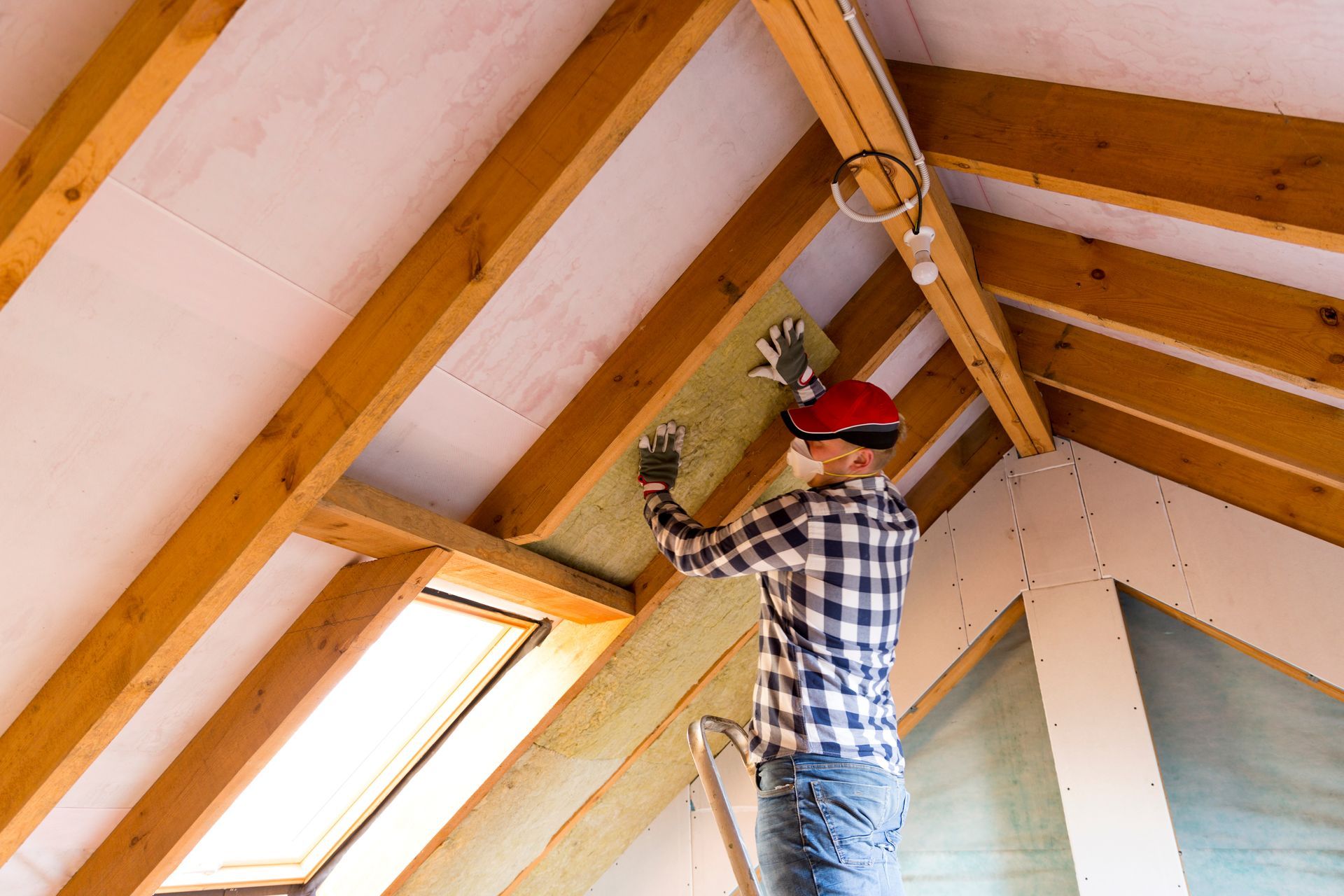 Ouvrier installant de l'isolant dans un plafond de grenier incliné à l'aide d'une perceuse et d'un casque de chantier rouge.