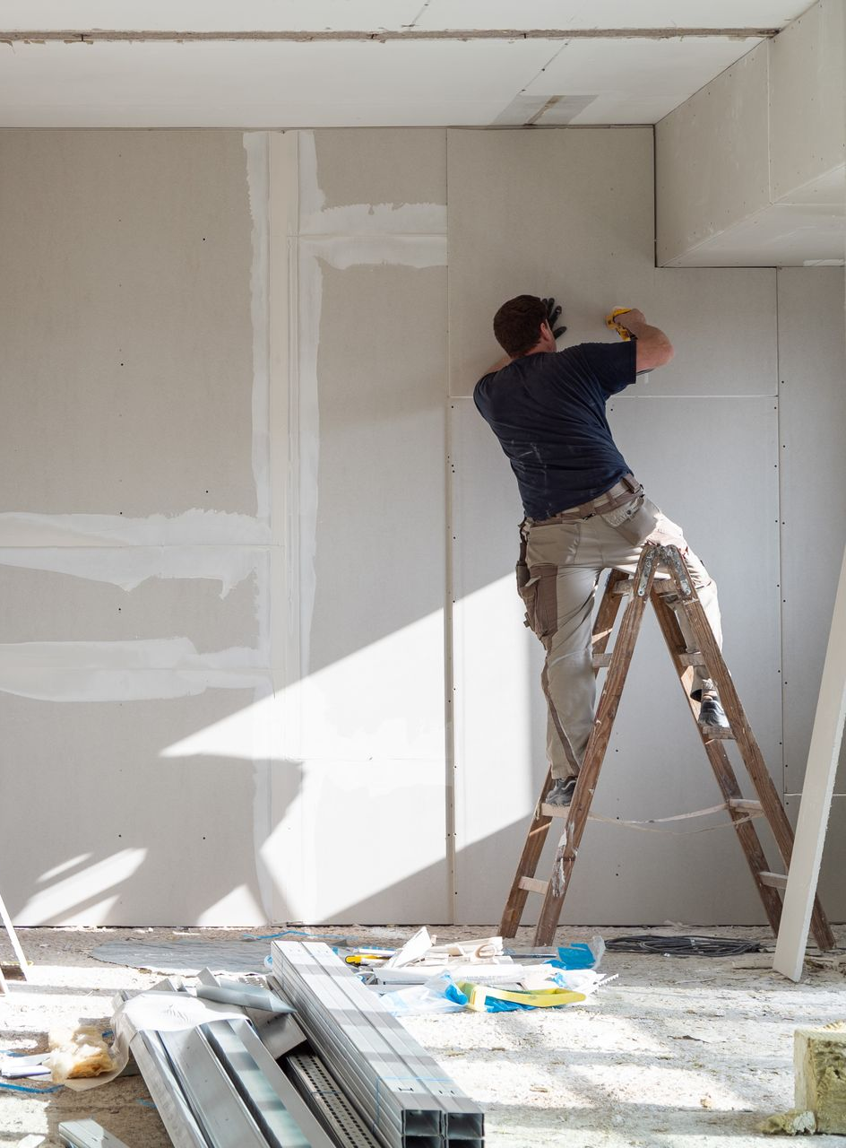 Une personne sur une échelle jointoyait des plaques de plâtre dans une pièce lumineuse en rénovation.