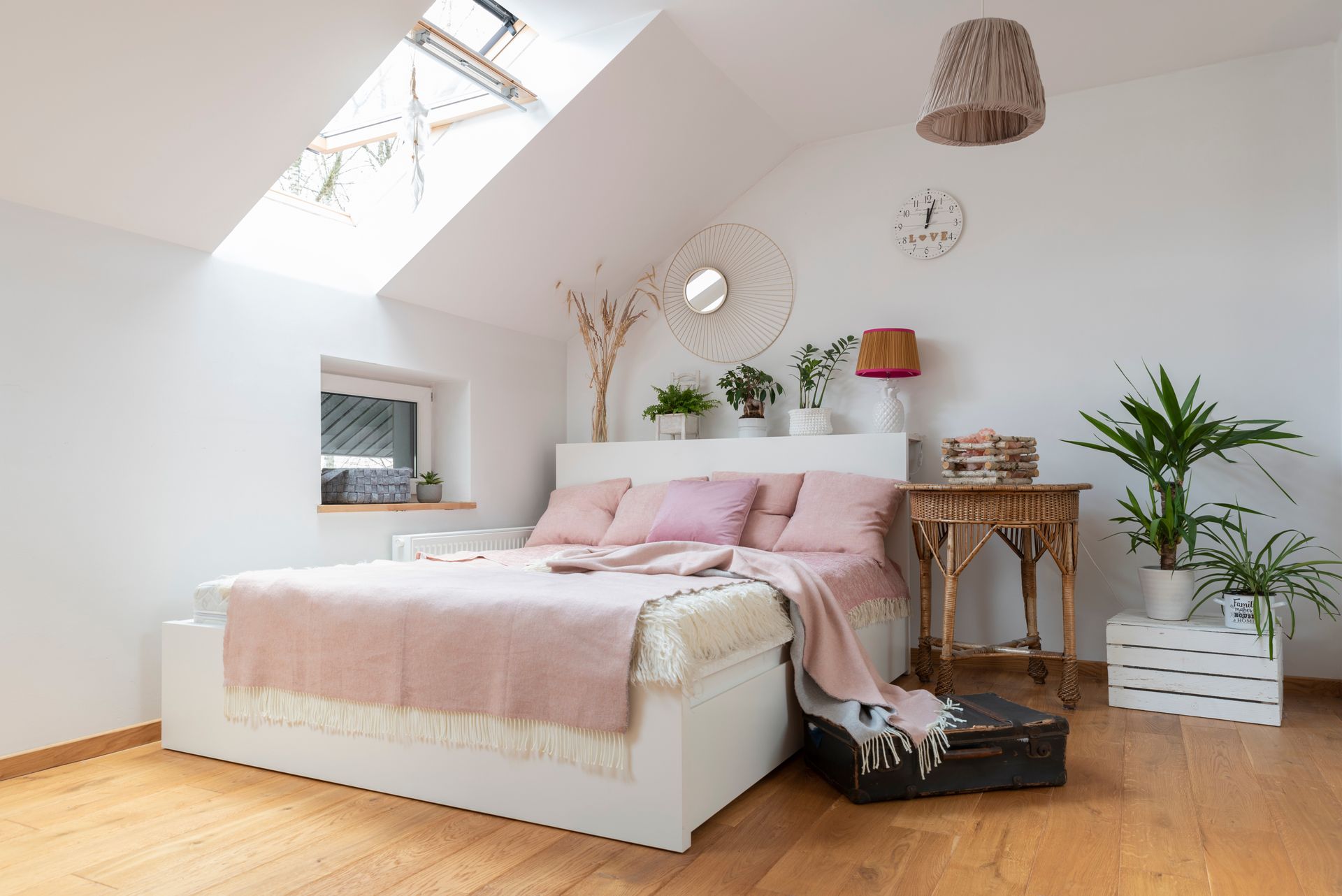 Chambre mansardée lumineuse avec literie rose, puits de lumière, plantes et parquet.
