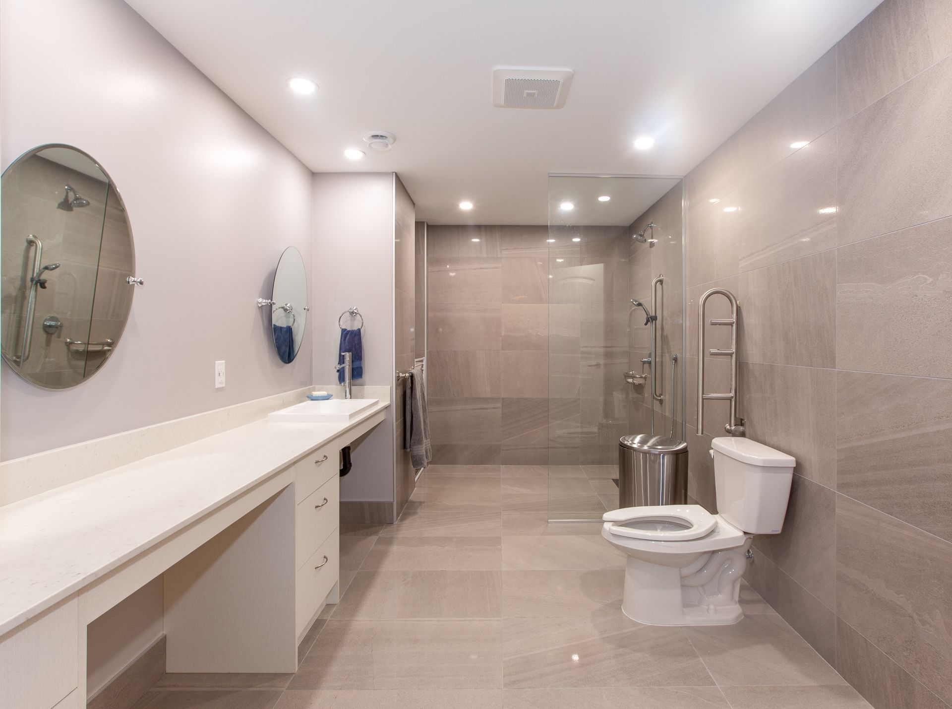 Salle de bain moderne et accessible avec lavabos, barres d'appui, toilettes et douche à l'italienne carrelée de beige.
