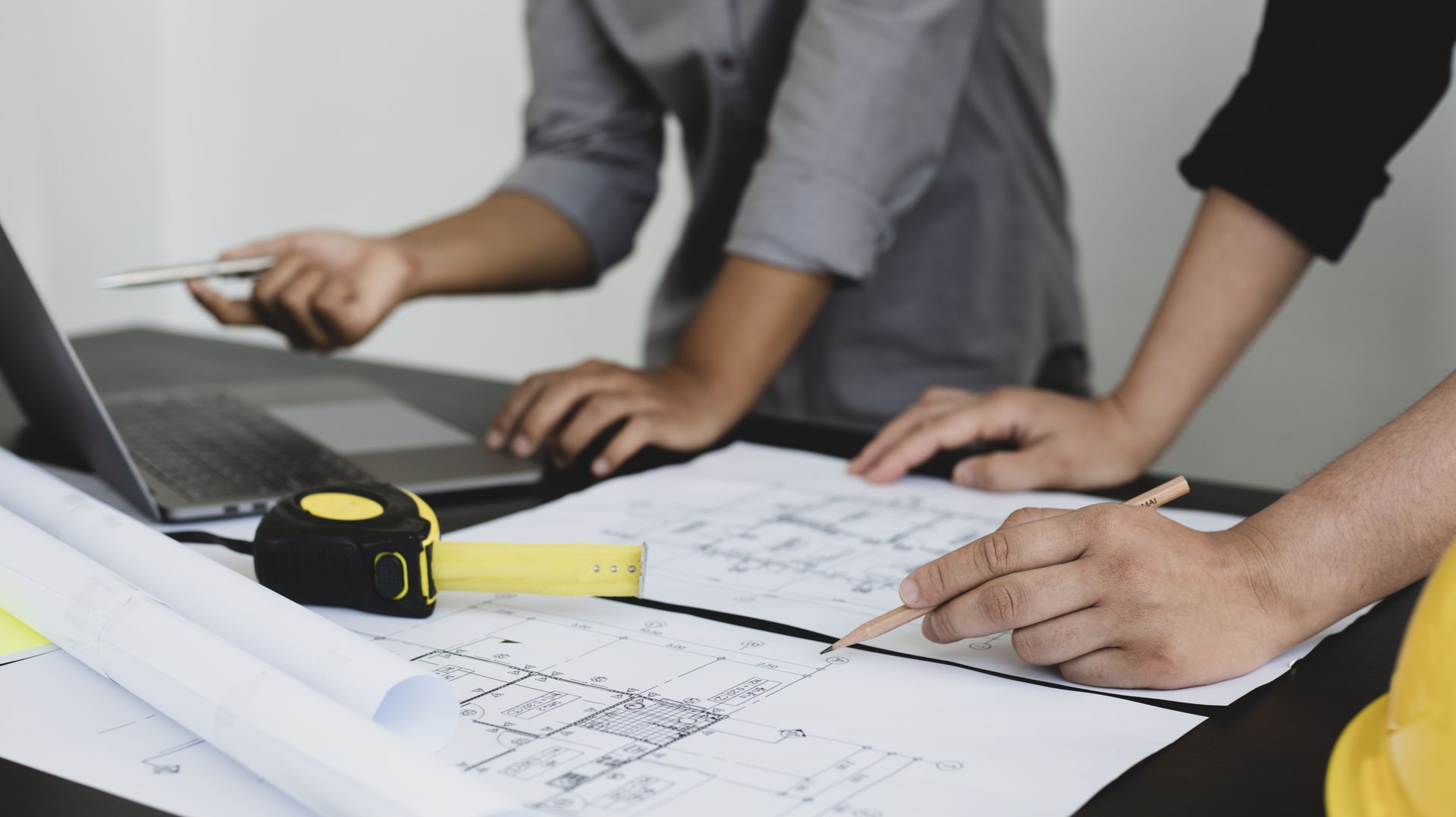 Des mains examinent un plan à côté d'un ordinateur portable, avec un mètre ruban et un crayon sur le bureau.