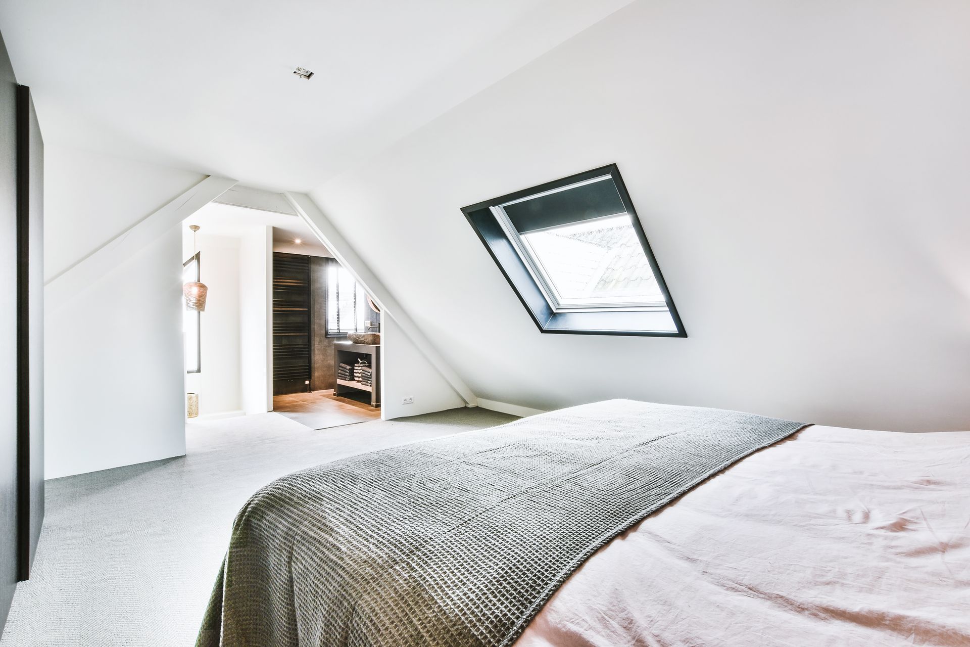 Chambre blanche lumineuse avec un lit et une fenêtre de toit inclinée sous un plafond en pente.