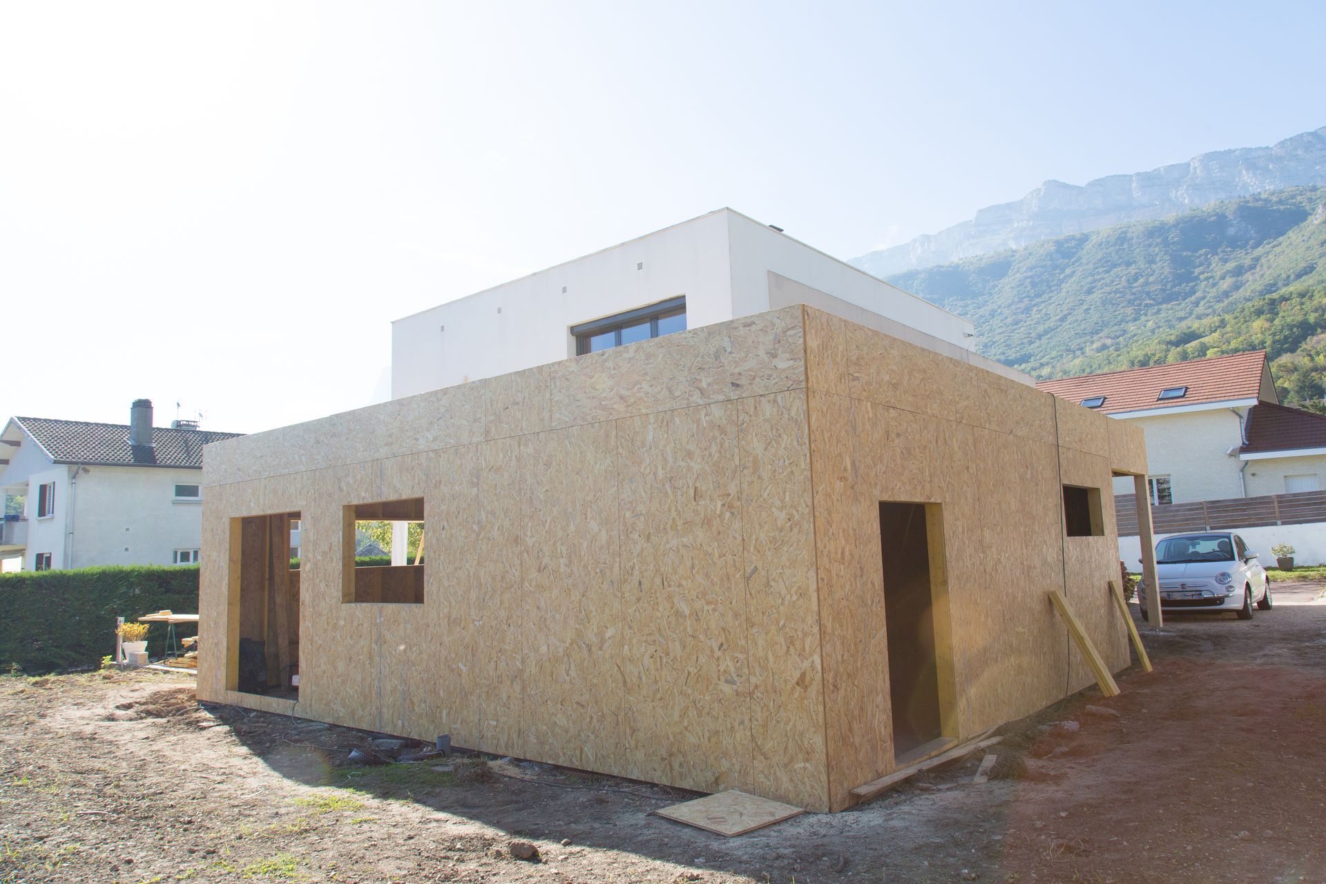 Maison moderne partiellement construite avec extérieur en contreplaqué dans une zone rurale ensoleillée, montagnes en arrière-plan.