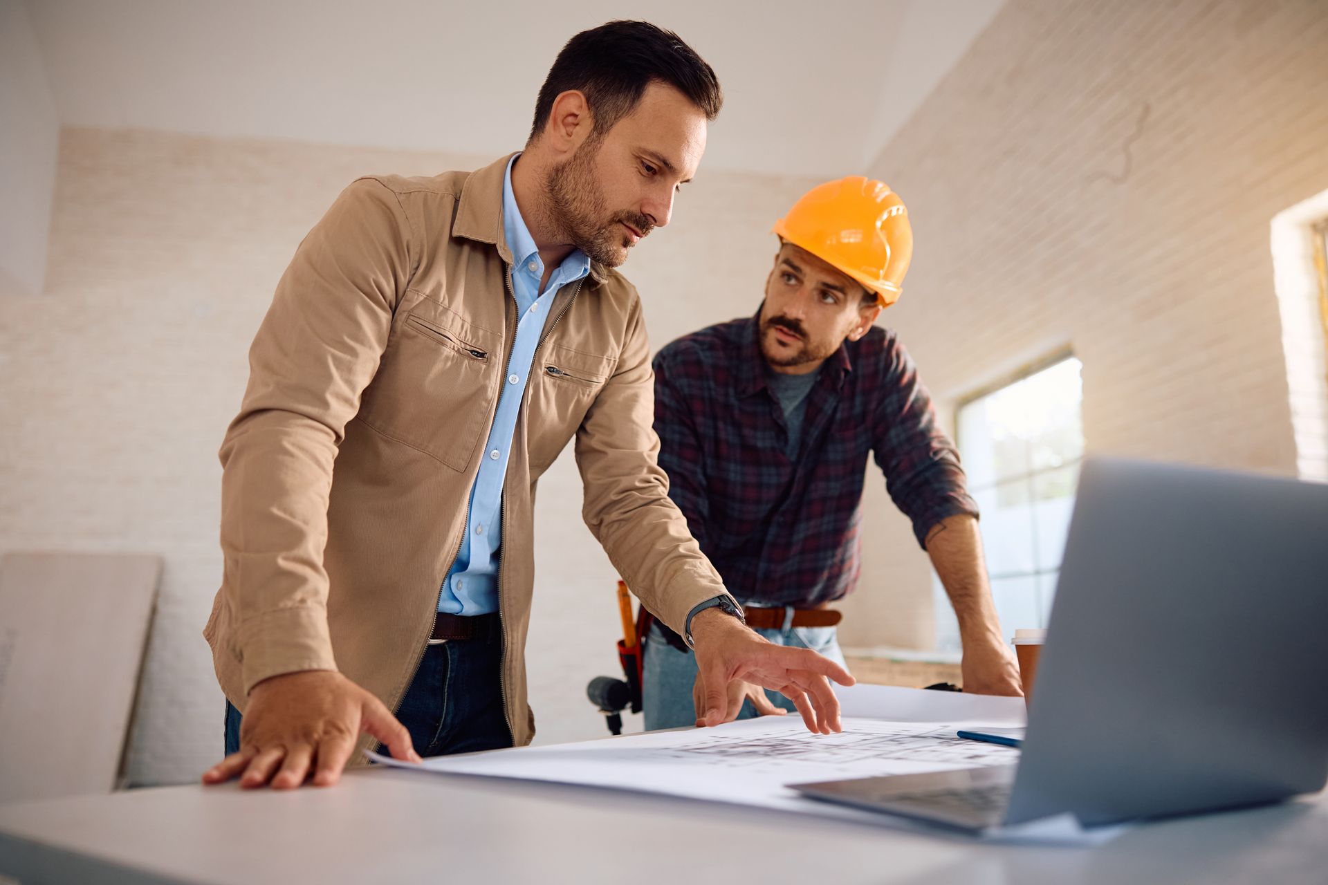 Deux ouvriers examinent des plans à un bureau, munis d'un ordinateur portable et d'un casque de chantier, dans un bureau lumineux.