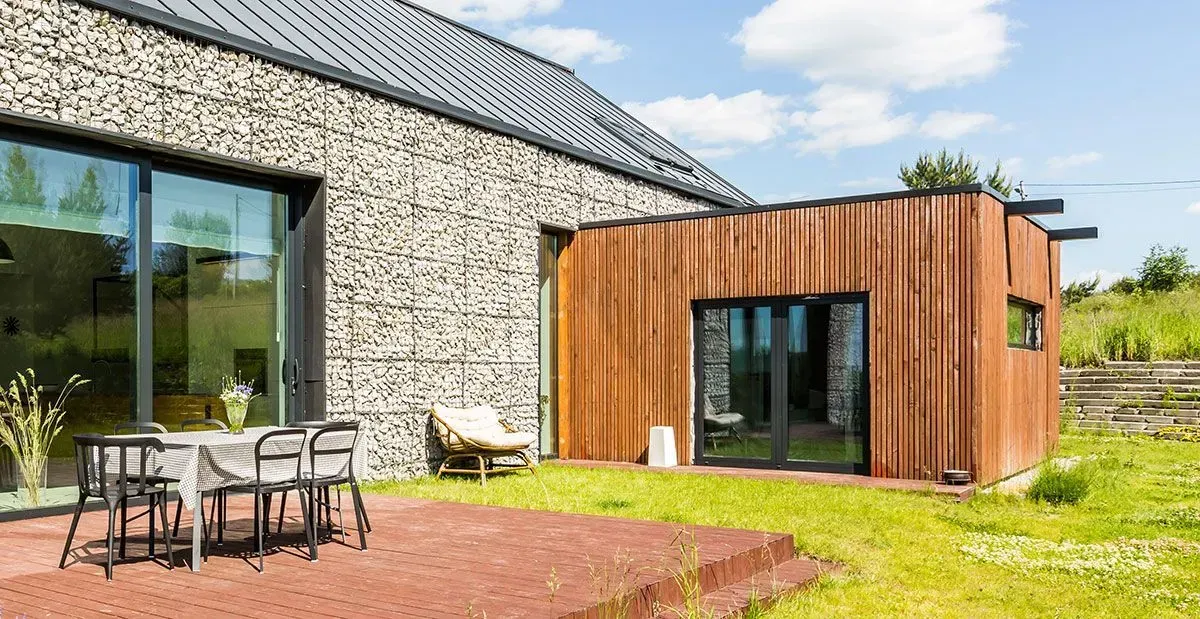 Maison en pierre avec portes vitrées et une petite extension en bois jouxtant une table et des chaises de patio.