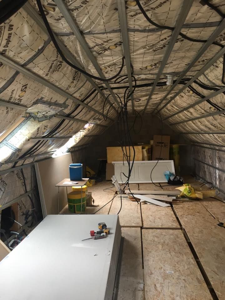 Atelier isolé dans les combles, avec poutres apparentes, planches, outils, câbles et une armoire blanche contre le mur du fond.
