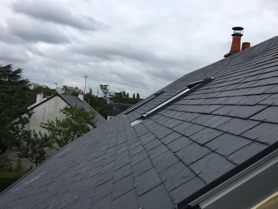 Toit en tuiles gris foncé avec puits de lumière et cheminée sous un ciel nuageux.