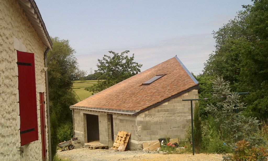 Ancienne dépendance en pierre avec un toit de tuiles en pente, jouxtant une maison, entourée d'arbres et de débris de jardin.
