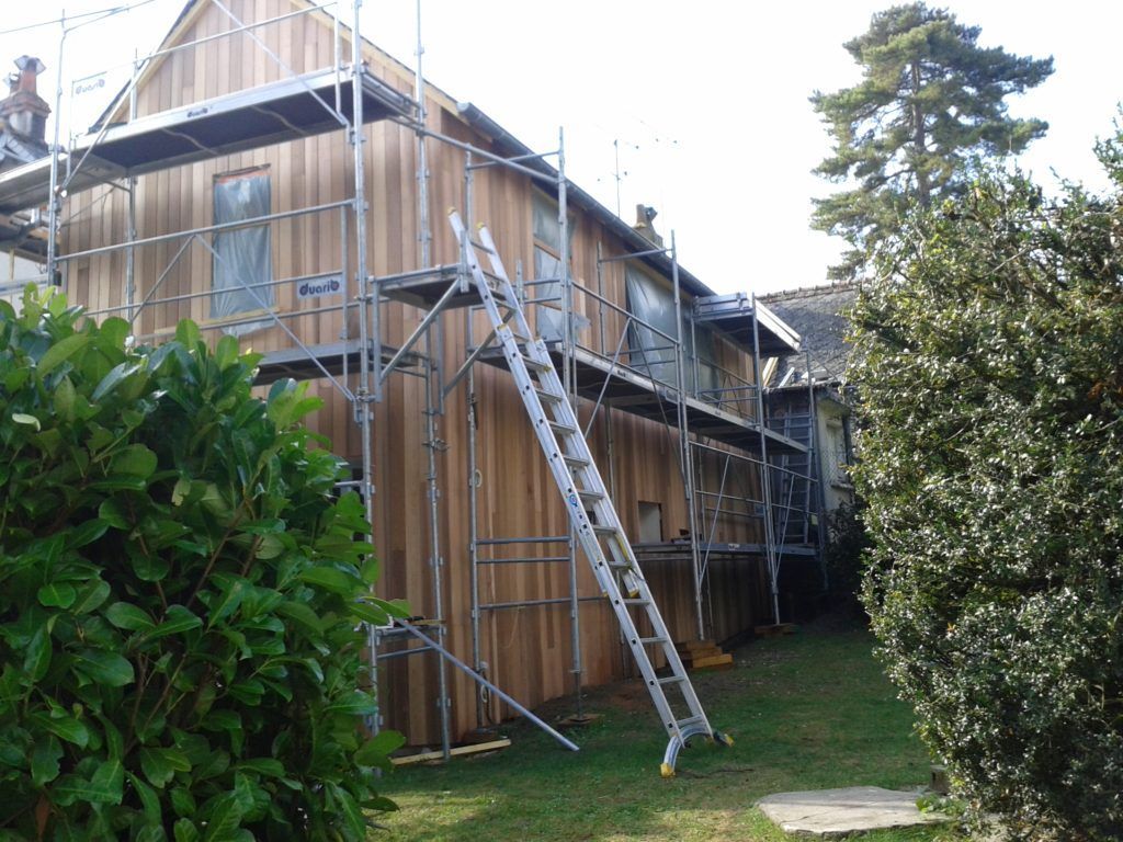 Échafaudages autour d'une maison en bois en construction, avec une échelle appuyée contre le mur.