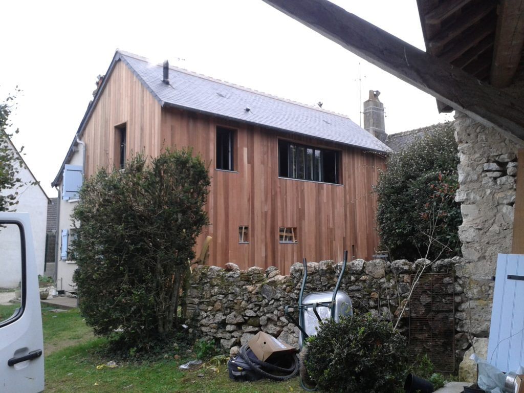 Maison en bois avec un mur de pierre et des buissons dans une cour rurale sous un ciel gris.