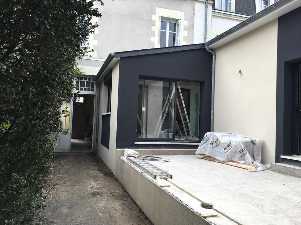 Extérieur d'une maison rénovée avec une extension gris foncé, une allée et des matériaux de couverture sur une terrasse en béton.
