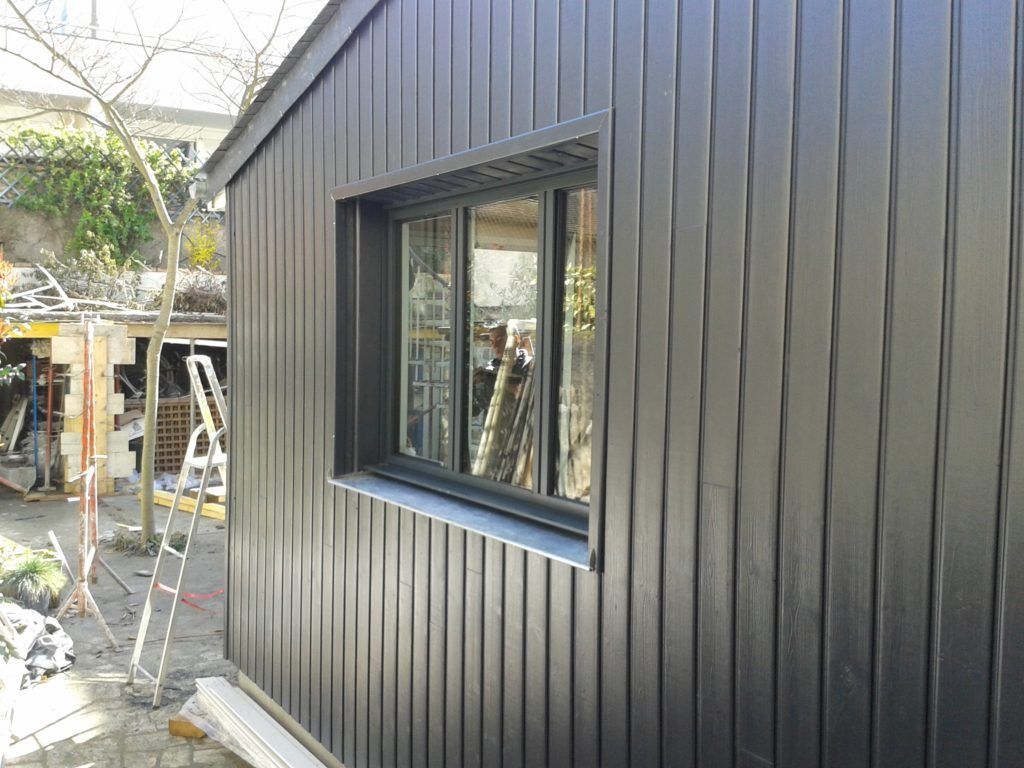 Façade d'un bâtiment aux parois noires, avec une seule fenêtre et des matériaux de construction à proximité.