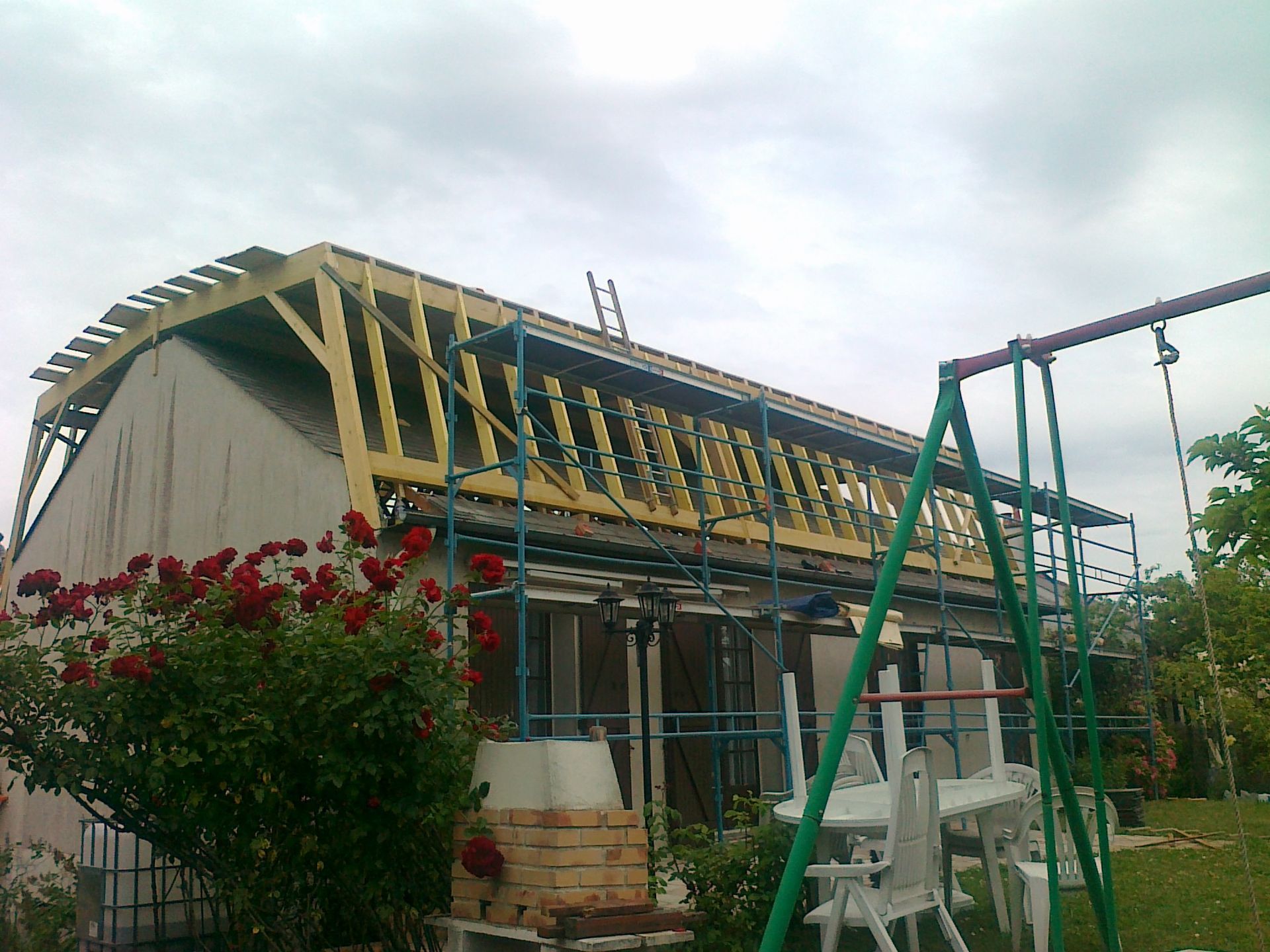 Maison en construction avec charpente de toit, échafaudages et échelles sous un ciel nuageux