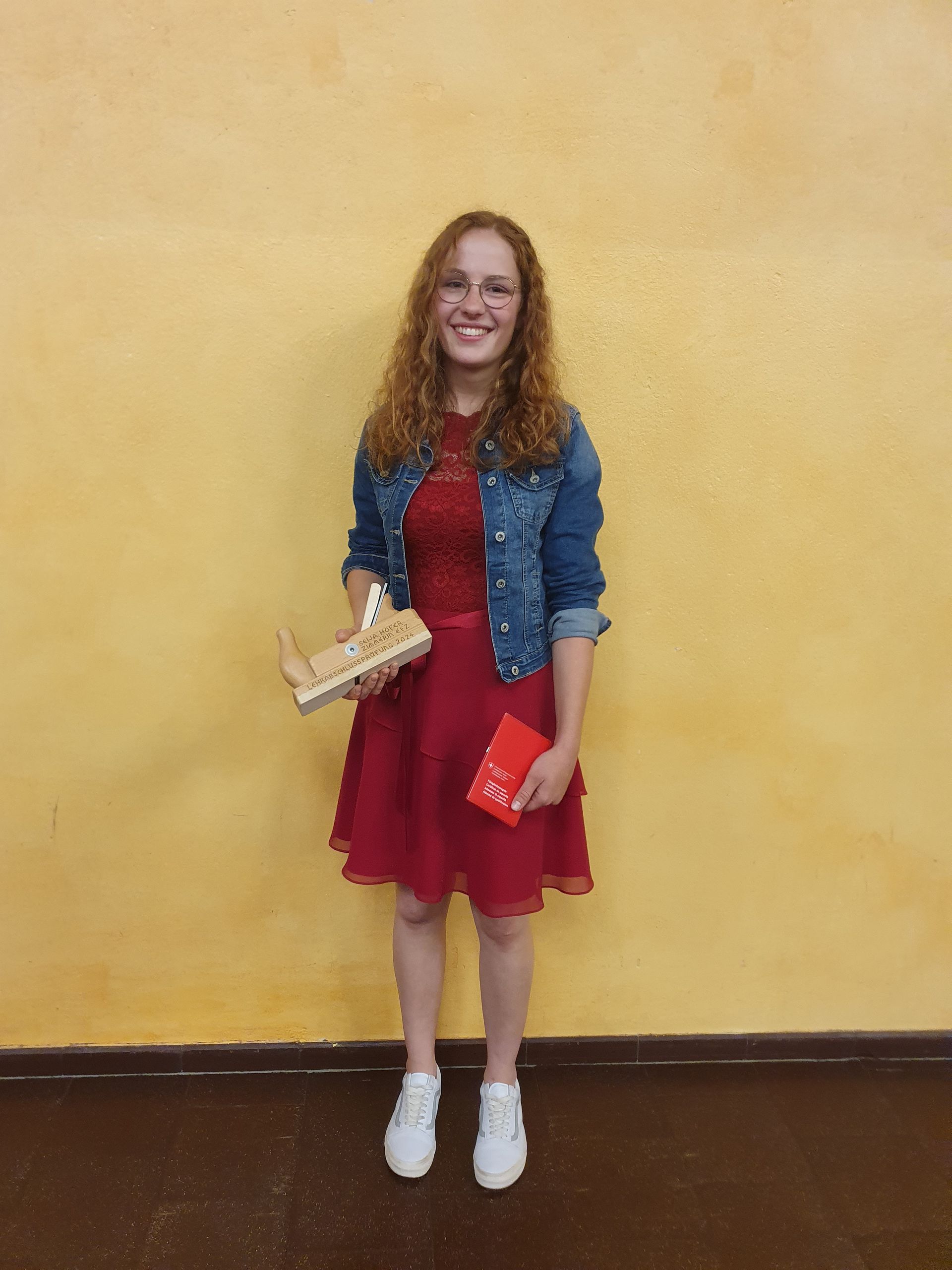 Eine Frau in einem roten Kleid und einer Jeansjacke steht vor einer gelben Wand und hält eine Trophäe in der Hand.