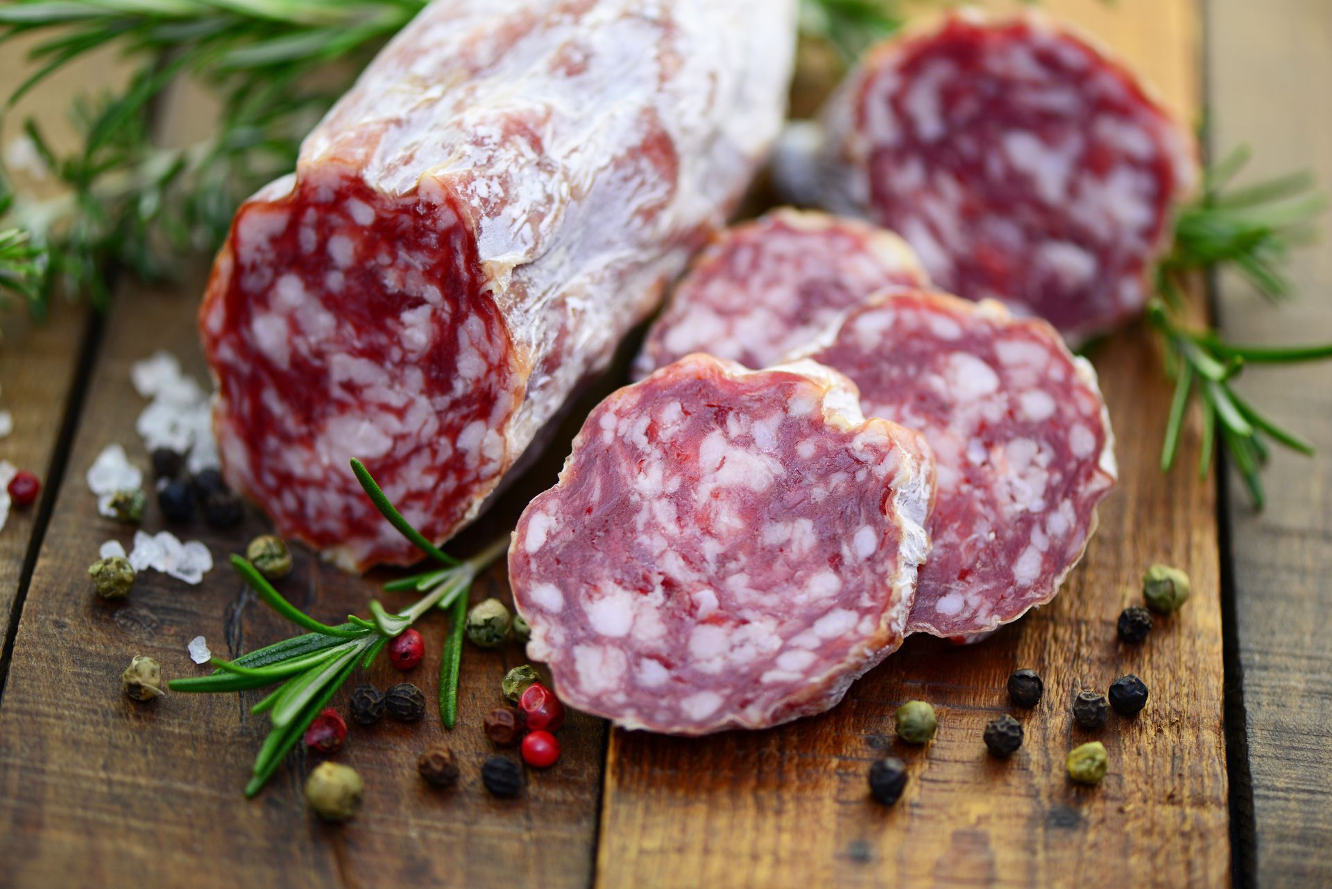 In Scheiben geschnittene Salami mit weißer Schwarte, Rosmarin und Pfefferkörnern auf einem Holzbrett.