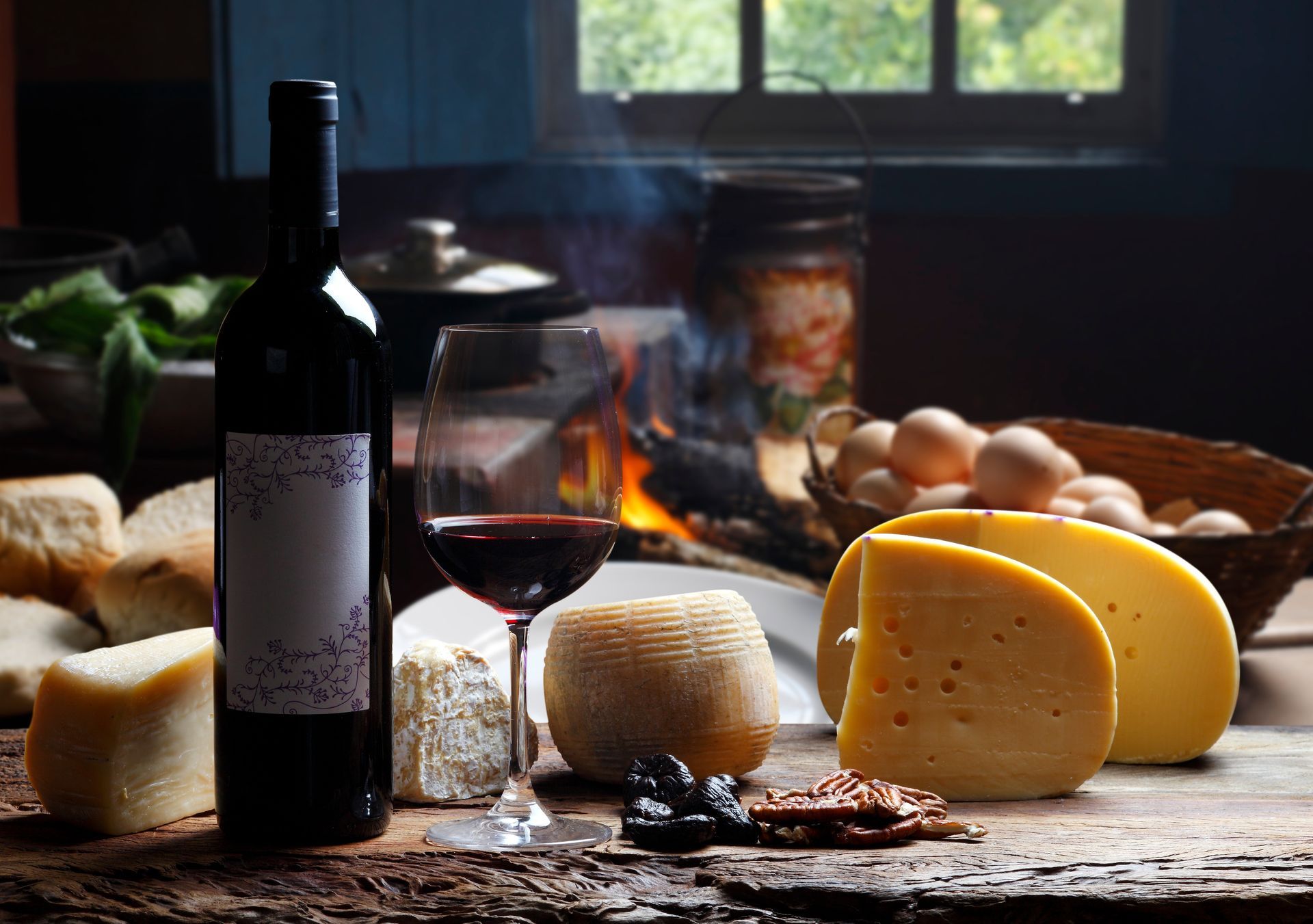 Eine Weinflasche, ein Glas Rotwein, Käse und Eier stehen auf einem Tisch vor einem Fenster und einem Kamin.