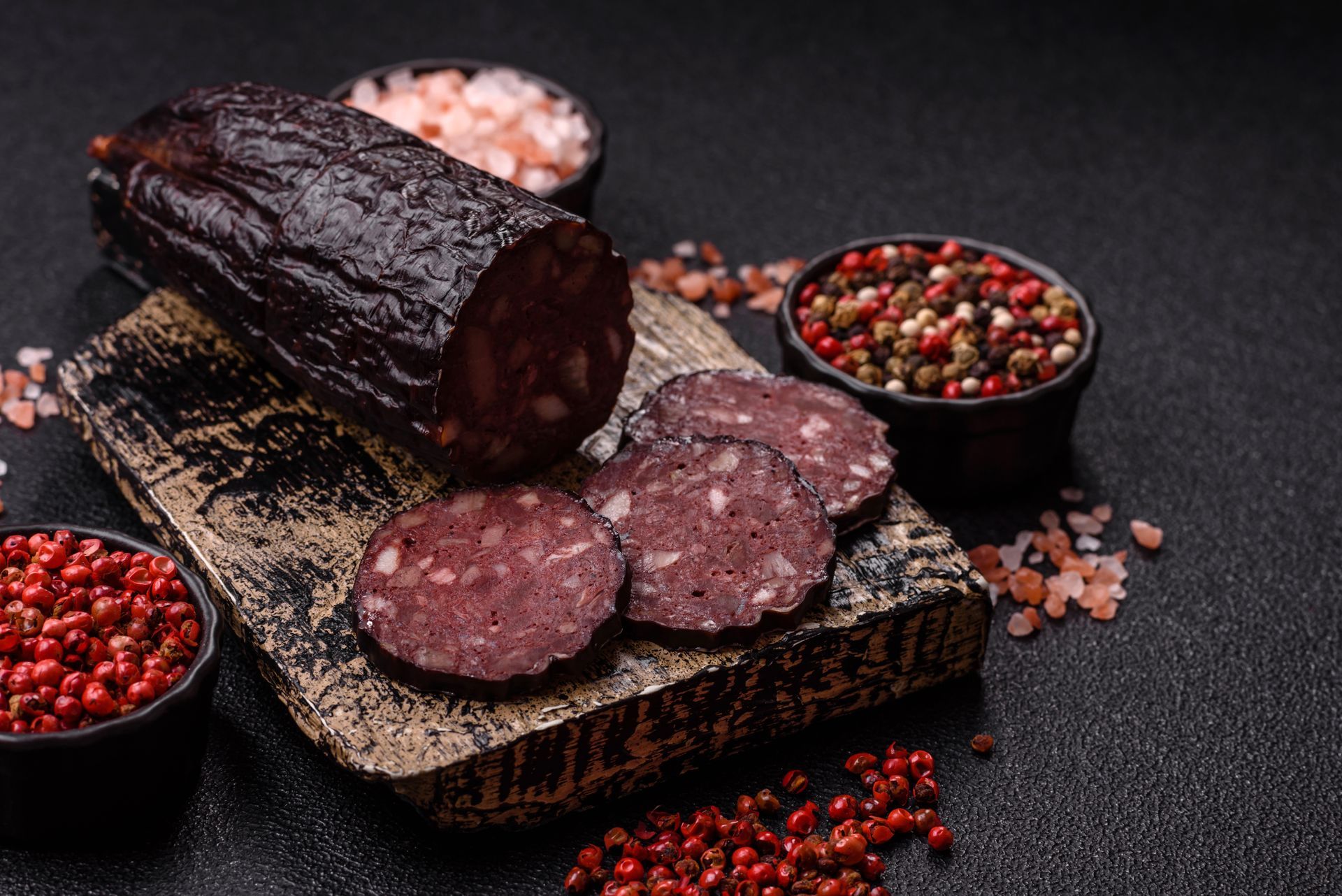 Aufgeschnittene, geräucherte Wurst auf einem Holzbrett mit Schälchen mit Pfefferkörnern und grobem Salz