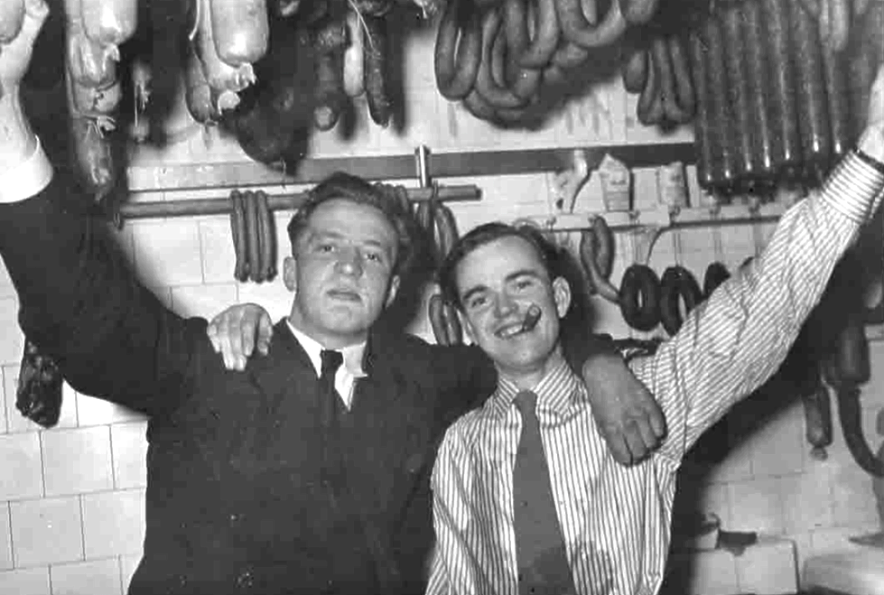 Zwei Männer in einer Metzgerei, die Arme erhoben, umgeben von hängenden Würsten und Fleischwaren. Einer mit einer Zigarre.