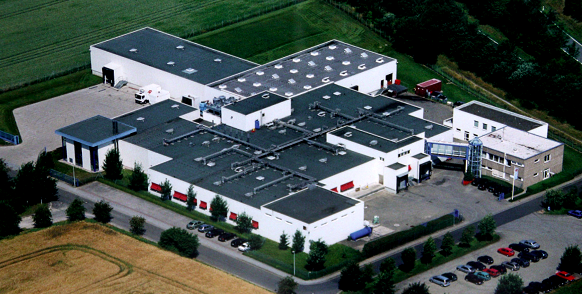 Luftaufnahme eines weißen Industriegebäudekomplexes mit dunklem Dach, umgeben von grünen Feldern.